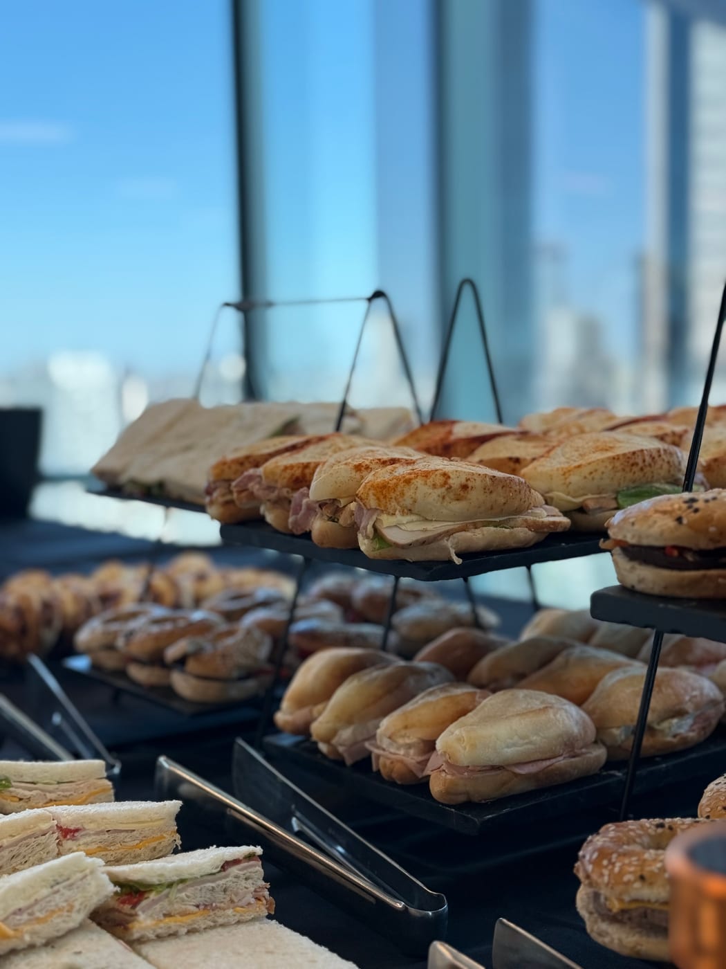 Buffet en niveles con sándwiches y pebetes frente a un ventanal con vista a la ciudad