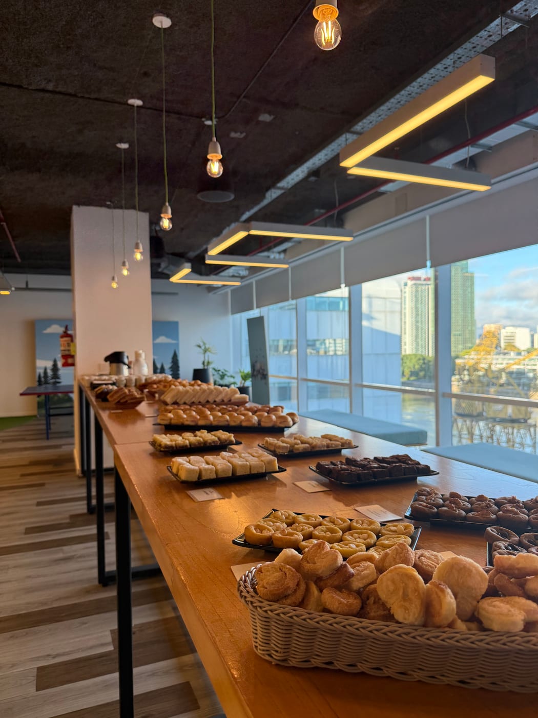 Mesa de desayuno corporativo con canastos de panes, medialunas y facturas junto a ventanales