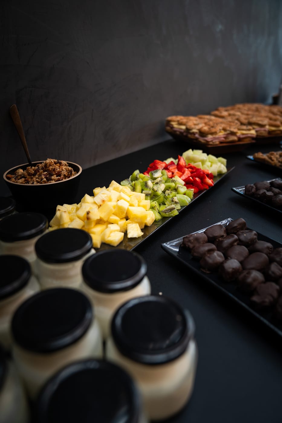 Mesa de desayuno con vasitos de yogur, bowl de granola con frutos secos, frutas frescas cortadas y sandwiches en un evento corporativo