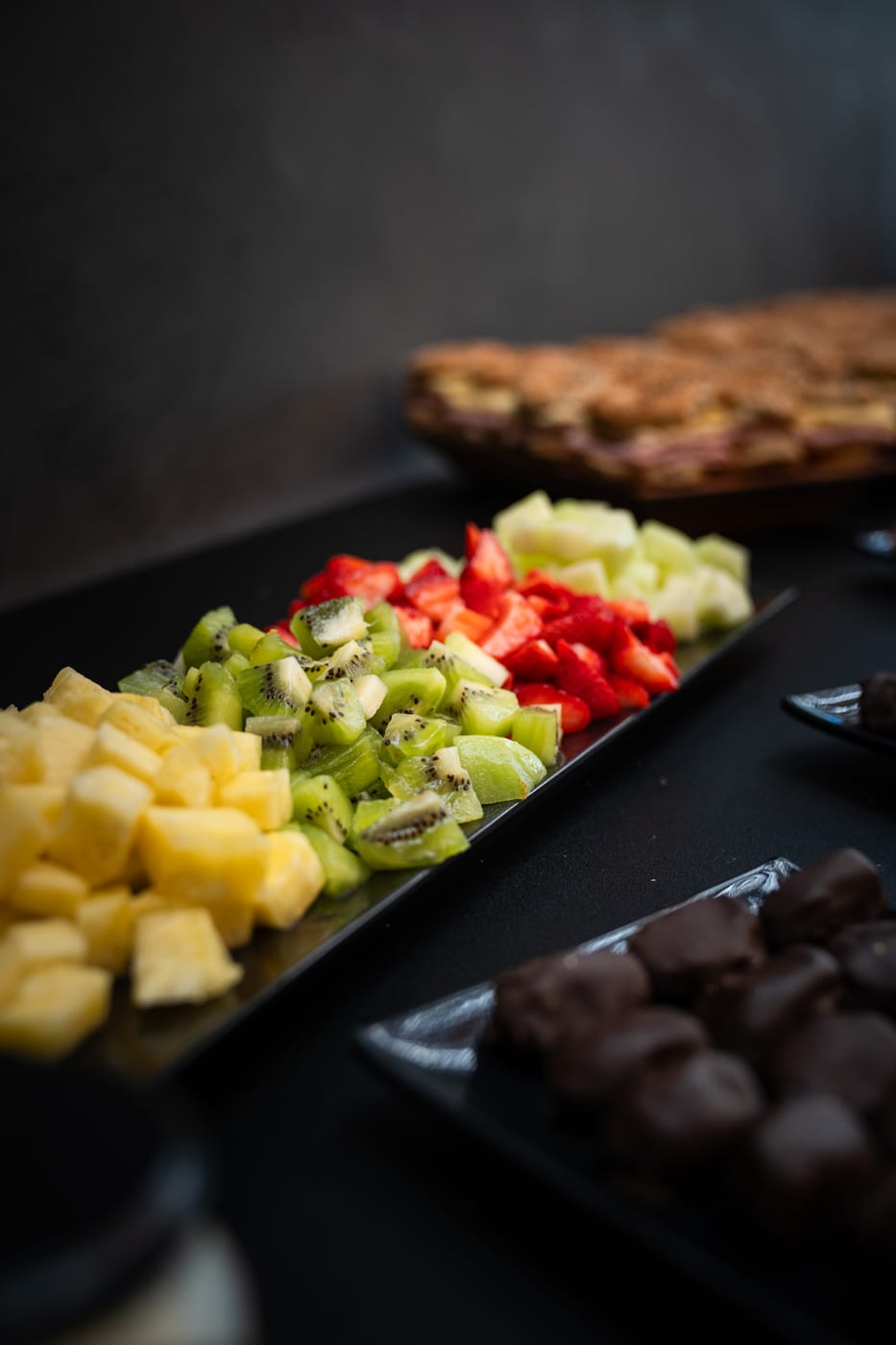 Bandeja de frutas frescas cortadas (ananas, kiwi, frutilla y manzana) junto a bombones de chocolate y sandwiches para un desayuno
