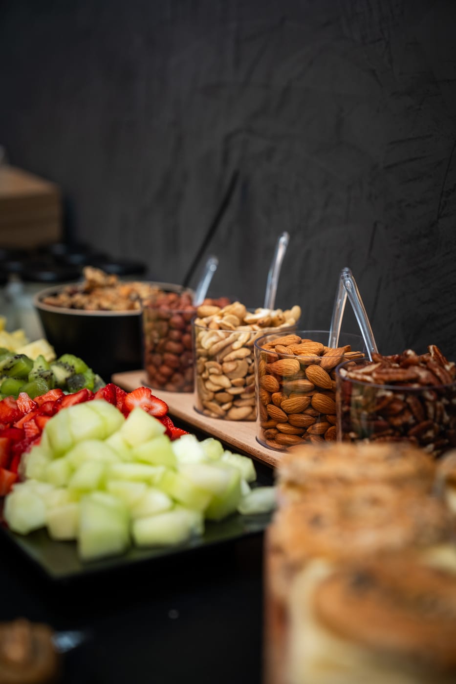 Detalle de vasitos con frutos secos (almendras, castañas de cajú, pecanas, avellanas), granola y frutas cortadas en una mesa de desayuno de catering
