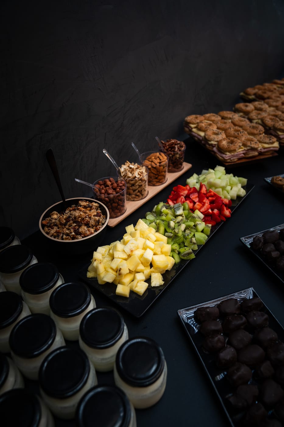 Vista general de una estacion de desayuno con frutas cortadas, granola, frutos secos, postres de chocolate y potes de yogur para un evento corporativo
