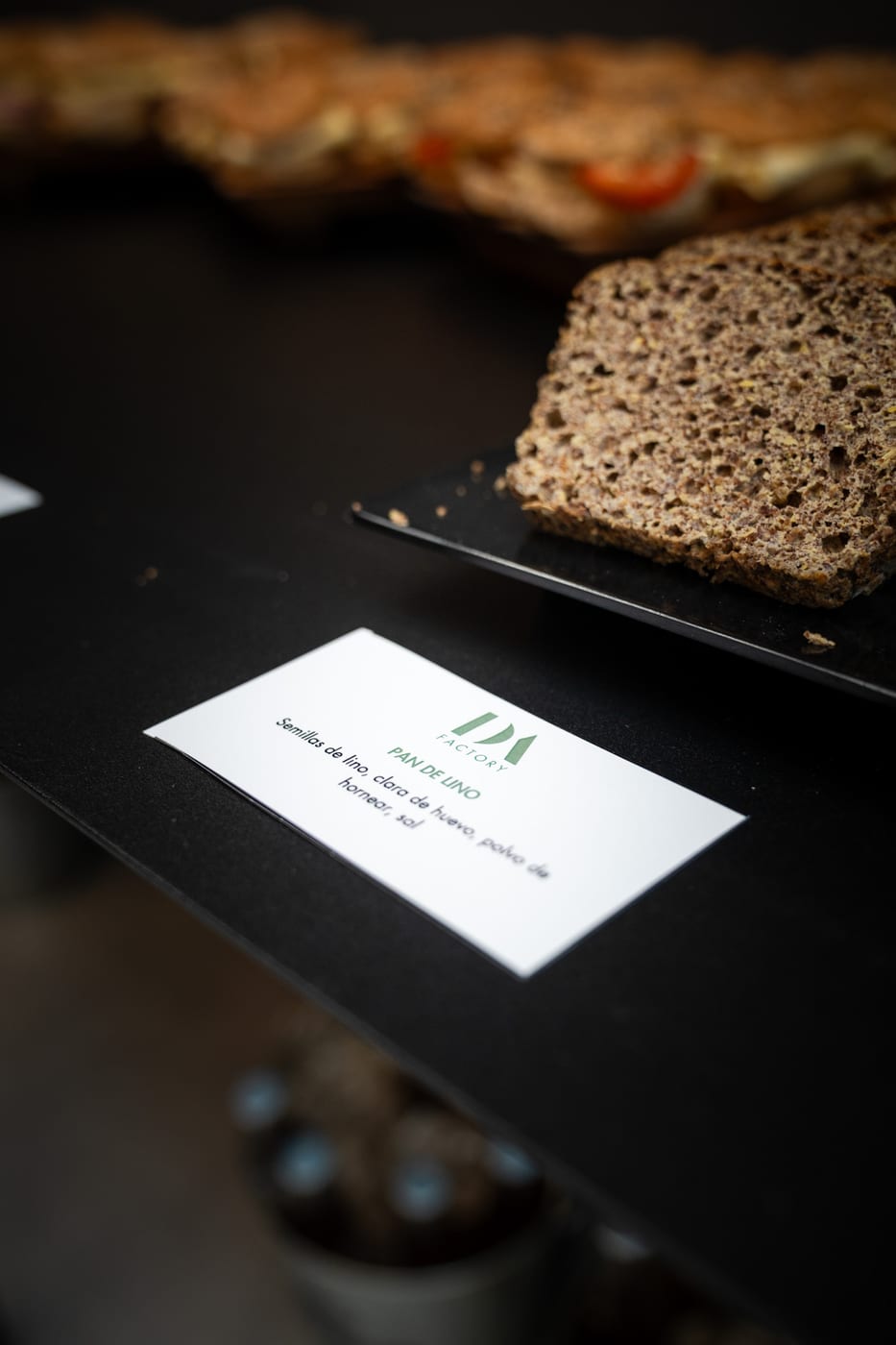 Pan de lino artesanal en rodajas con cartel de ingredientes de DA Factory, junto a sandwiches integrales, en un buffet de catering