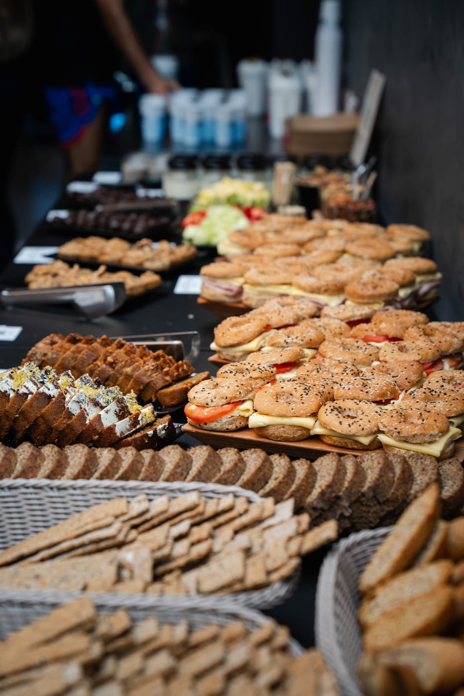 Buffet de sandwiches en pan de campo y bagels con queso y tomate, junto a canastas de panes integrales para un almuerzo corporativo