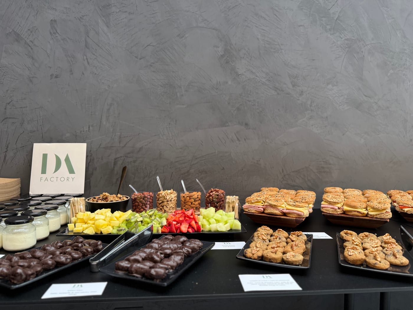 Mesa de coffee break corporativo con cartel DA Factory, vasitos de postre, frutos secos, brochettes de fruta, alfajores de chocolate, mini sandwiches en pan de sesamo y frutas frescas