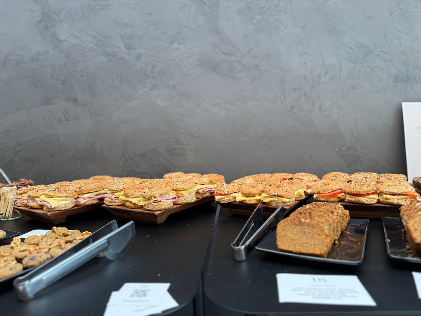 Bandejas de mini sandwiches en pan de sesamo con jamon, queso y tomate junto a budines caseros y cookies sobre mesa negra de evento