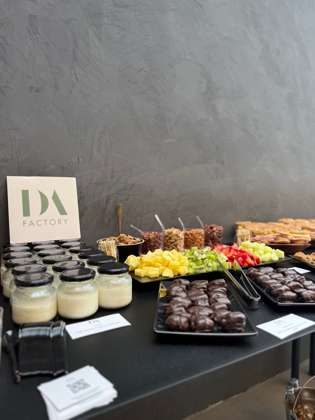 Coffee break con cartel DA Factory: frascos de postre tipo flan, brochettes de fruta, frutos secos, bombones de chocolate y mini pasteleria sobre mesa negra