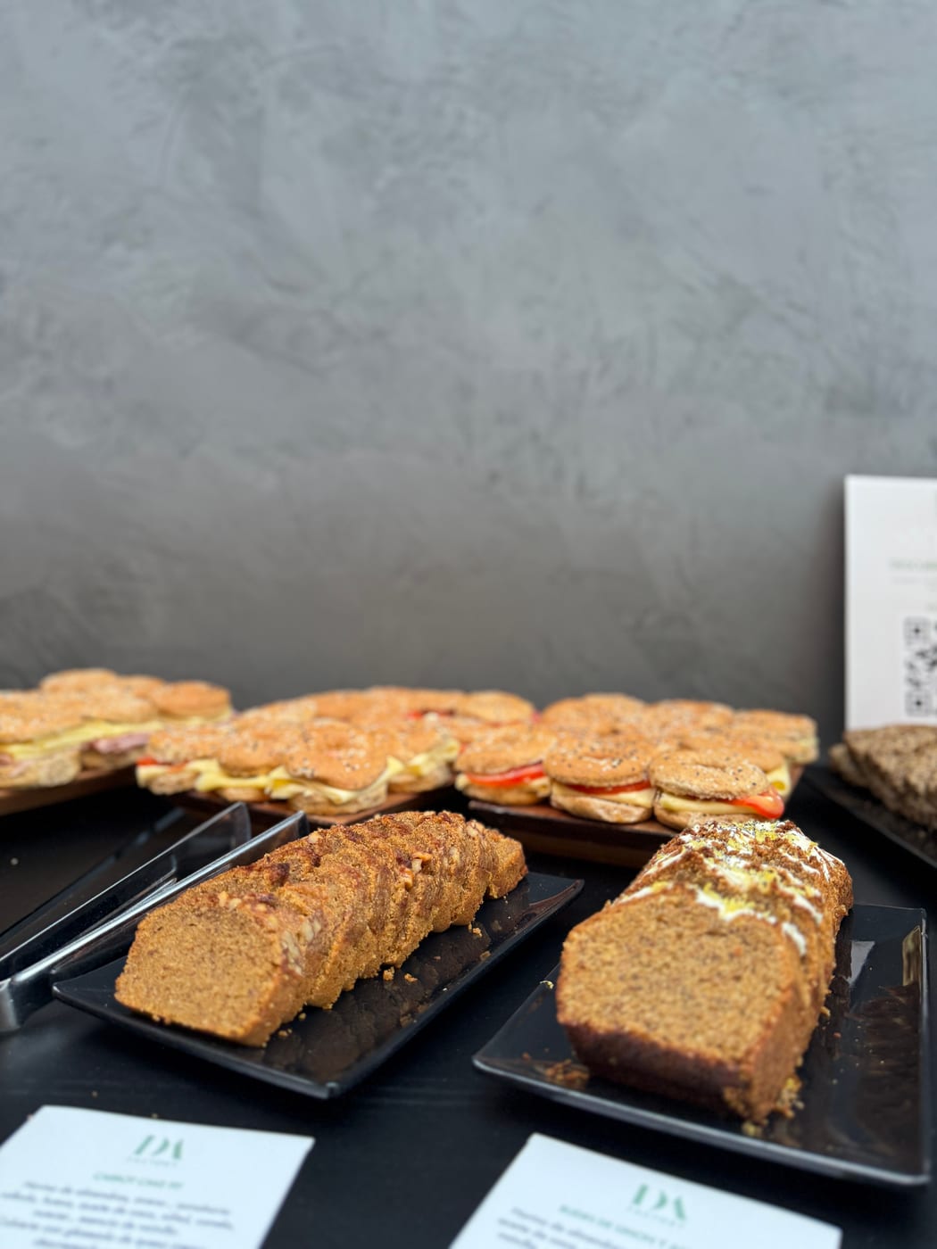 Budines caseros en rodajas sobre platos negros junto a bandejas de mini sandwiches en pan de sesamo para un servicio corporativo