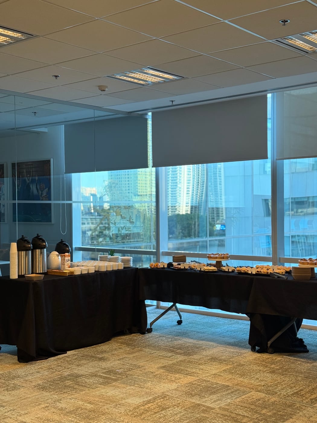 Montaje de desayuno corporativo en oficina con ventanal: termos de cafe, pilas de tazas, panificados y facturas sobre mesas vestidas de negro con vista a la ciudad