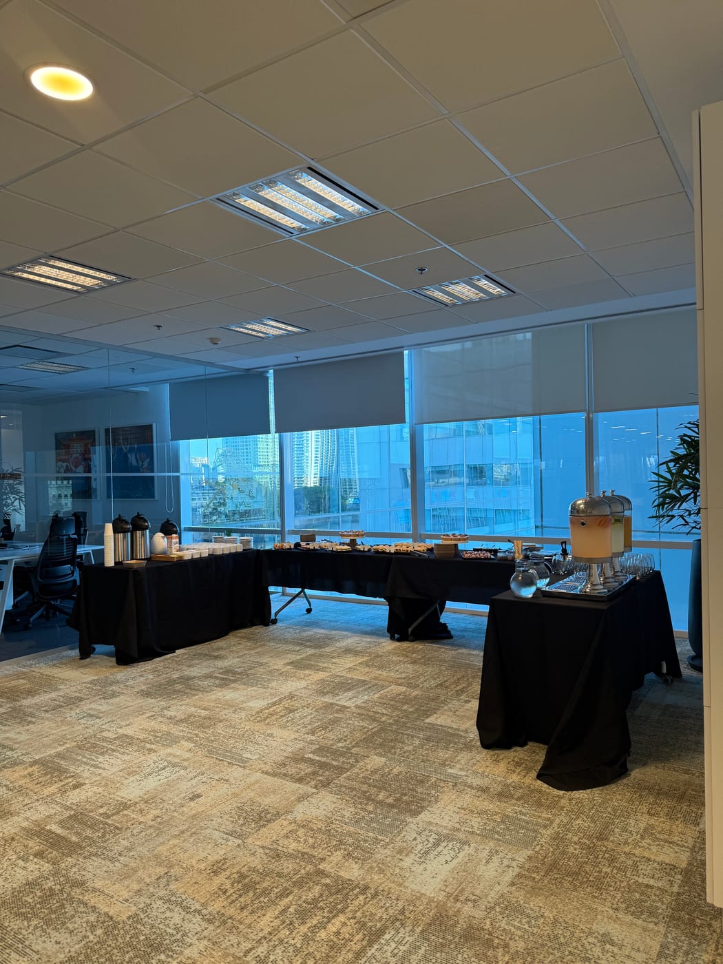 Montaje de coffee break corporativo con termos de cafe y bandejas sobre mesas vestidas en negro frente a ventanales de oficina