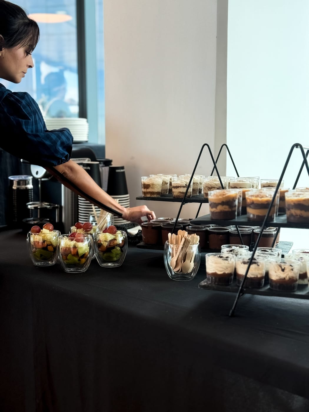 Mesa de coffee break con vasitos de postres tipo tiramisu y mousse de chocolate en pisos, ensalada de frutas en vasos y maquina de cafe, montaje para evento corporativo