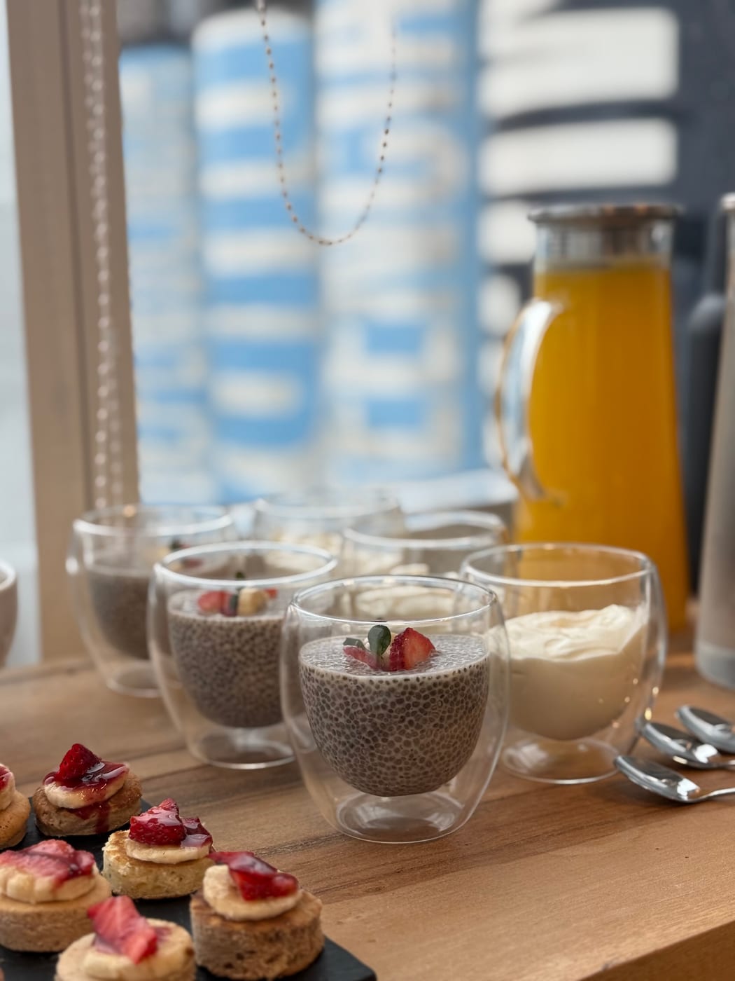 Vasos de pudding de chia con frutillas, jarra de jugo de naranja, crema batida y tostaditas con banana y mermelada de frutilla, mesa de desayuno saludable