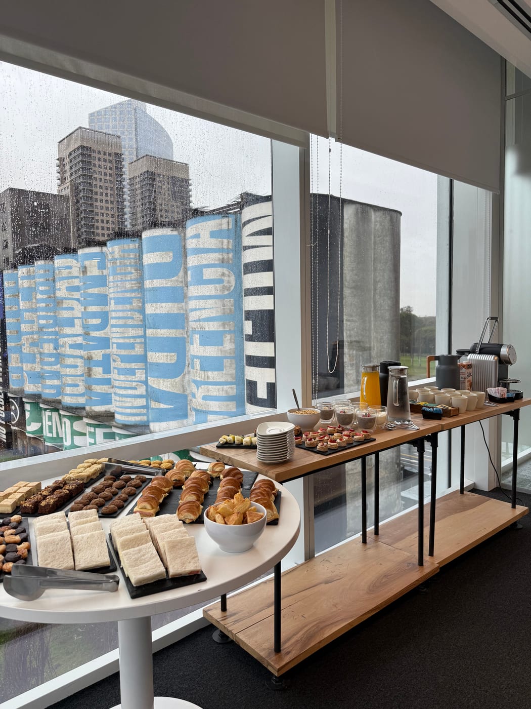 Estacion de desayuno tipo buffet con panificados, medialunas, tostadas y dispensadores de jugo sobre barra de madera junto a ventanal