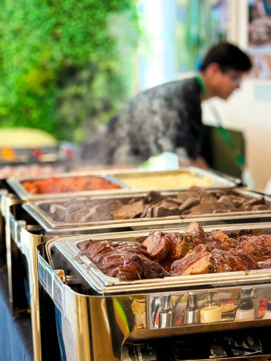 Buffet caliente con bandejas de carne braseada al vino, cerdo y guarniciones humeantes en chafing dishes de acero, con personal de servicio detras