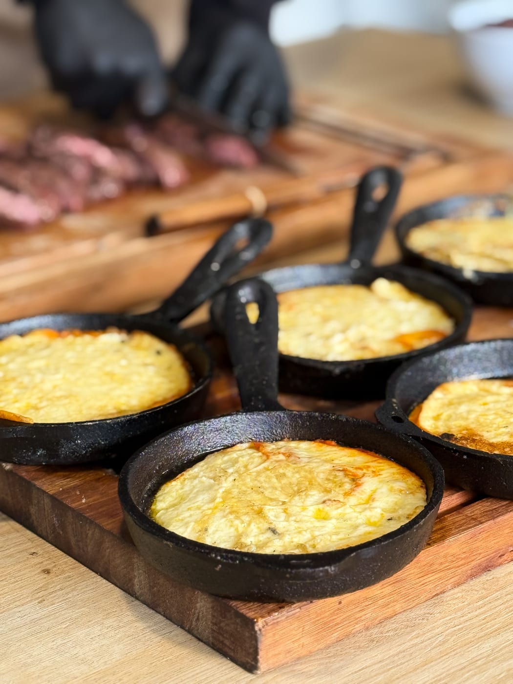 Cazuelas individuales de provoleta gratinada en sartenes de hierro sobre tabla de madera, con cortes de carne al fondo, en un almuerzo corporativo