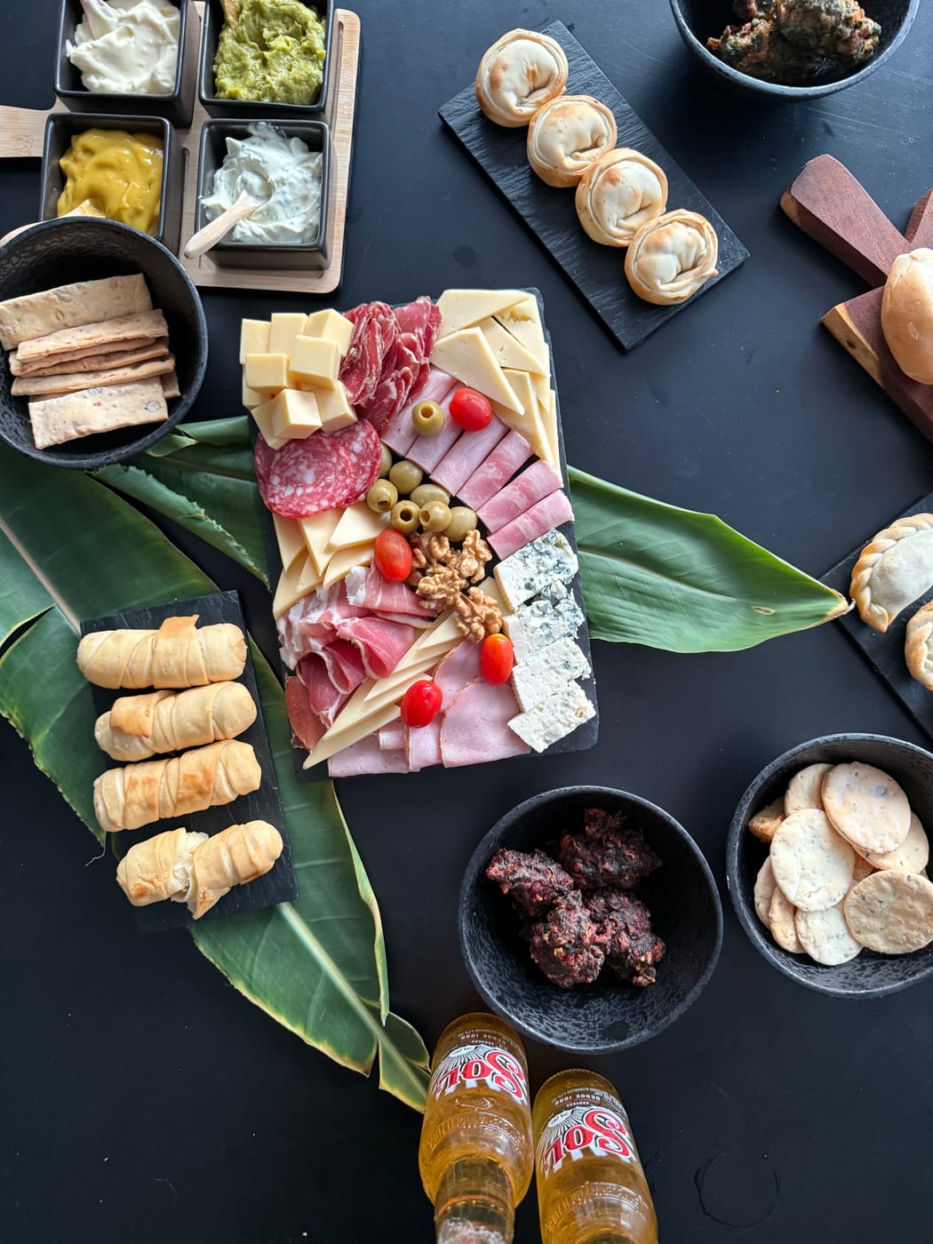 Tabla de fiambres y quesos con jamon, salame, aceitunas y nueces, rodeada de mini pastelitos, cuadraditos, dips y cervezas para una recepcion