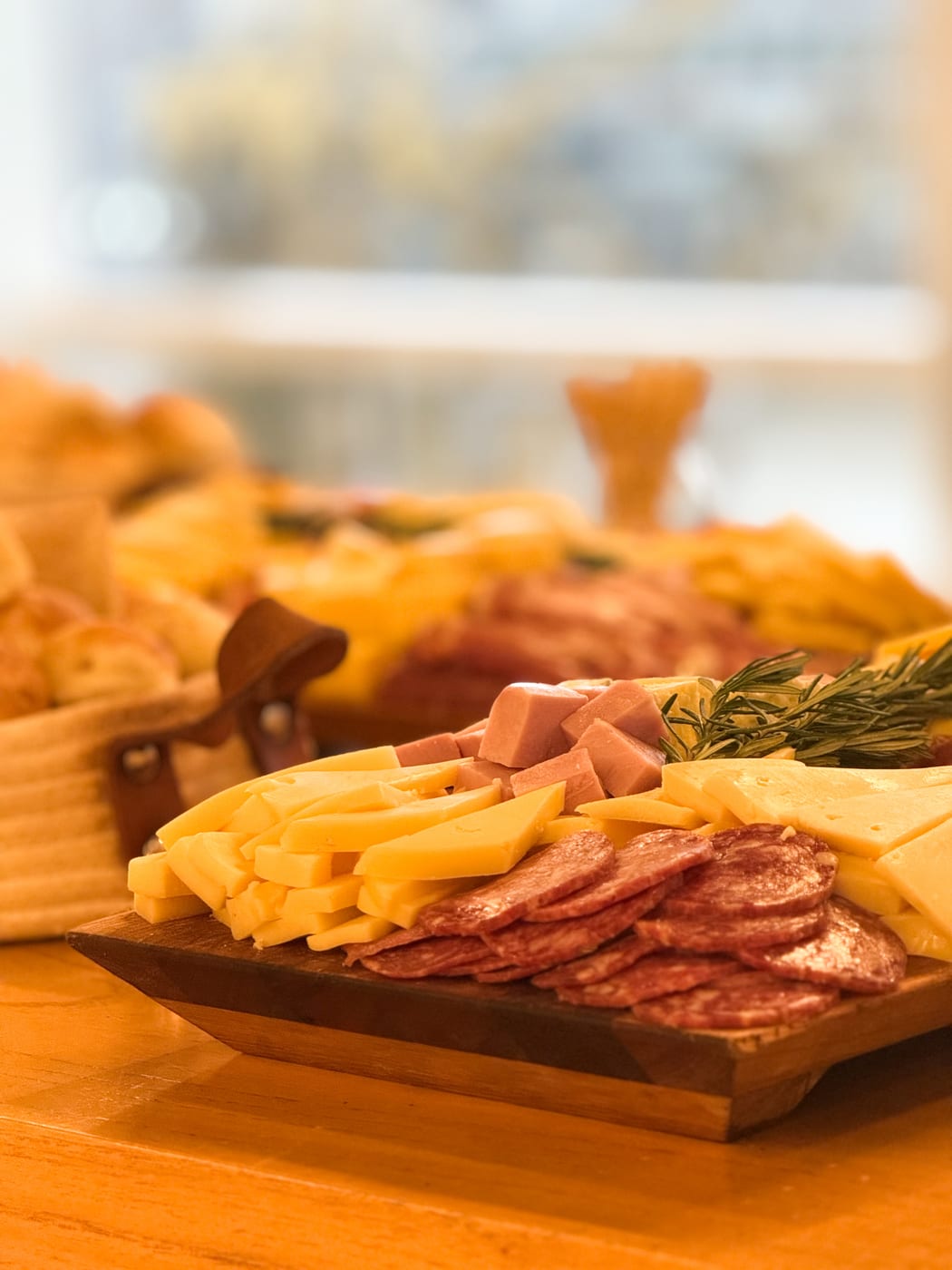 Tabla de quesos y fiambres con salame, jamon en cubos, quesos en cuna y feta, decorada con romero, junto a un canasto de panes
