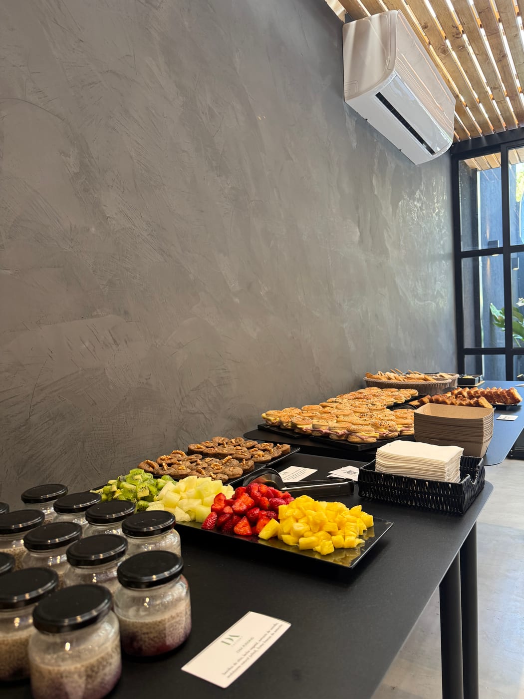 Estacion de coffee break con bandejas de galletas, frutas frescas cortadas (mango, frutillas), granola y jugos individuales montada sobre una barra