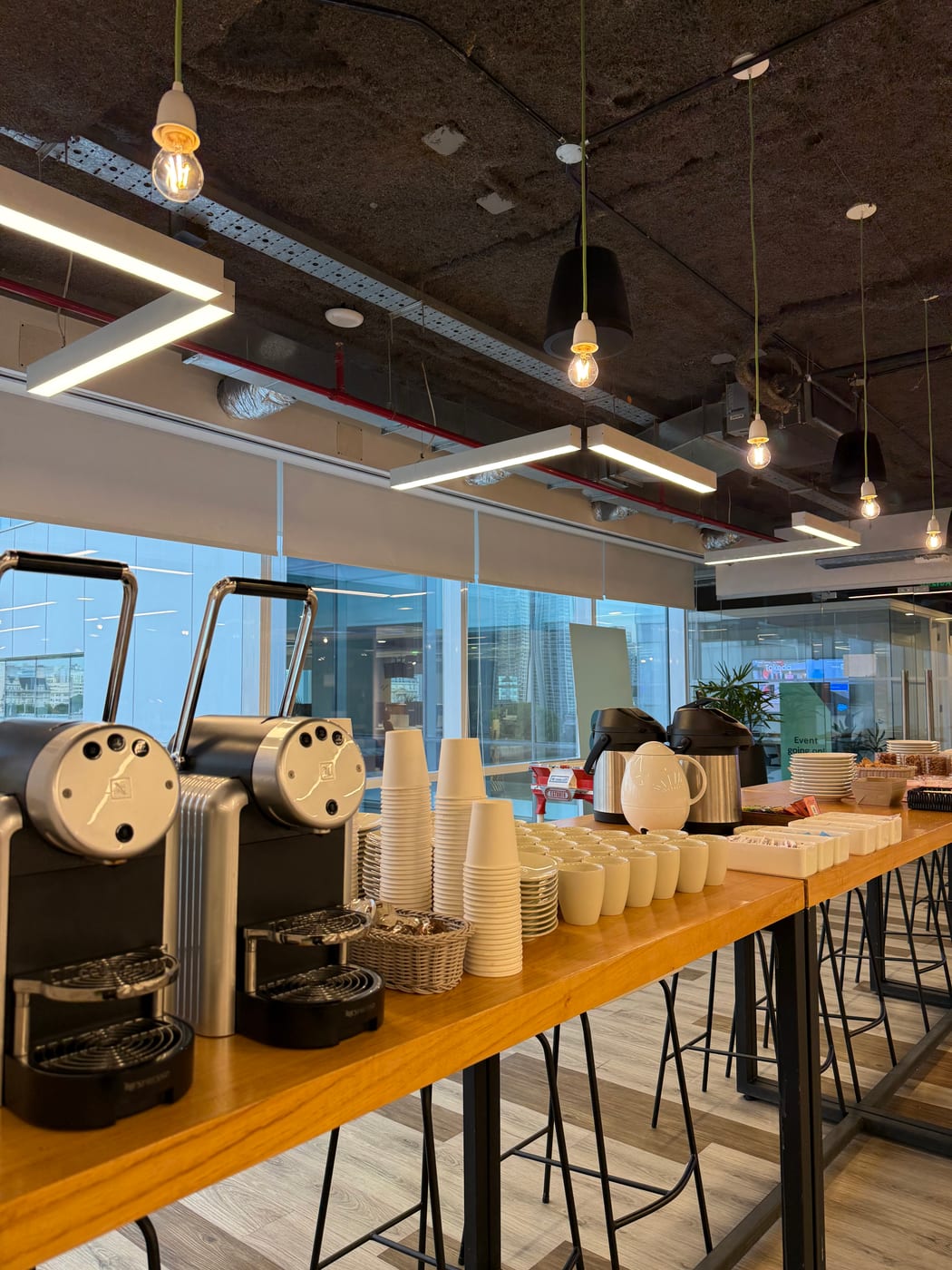 Estacion de cafe con maquinas expendedoras, torres de vasos y tazas montada sobre una barra de madera en un salon de eventos